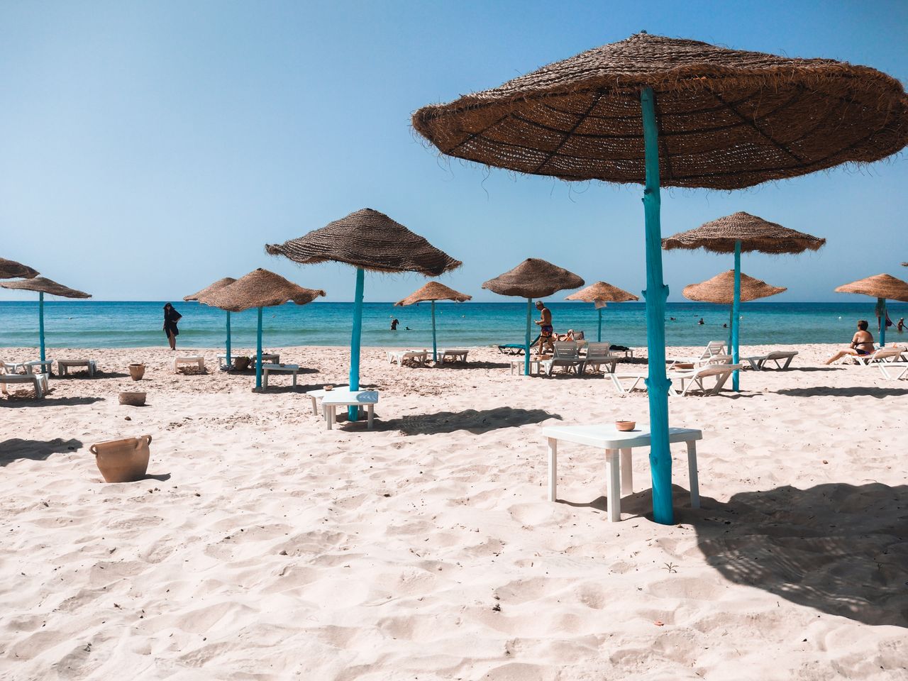 Beach scene with straw umbrellas, white sand, and clear blue sea under a bright summer sky.